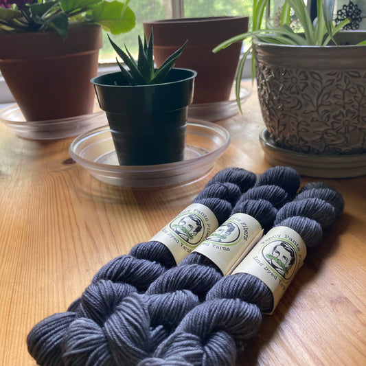 Three mini skeins of blue-grey yarn with labels on a wooden surface, surrounded by potted plants.
