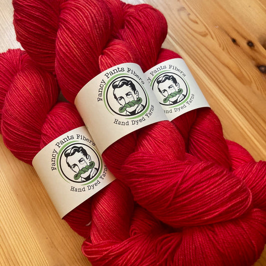 Red yarn skeins with 'Fancy Pants Fibers' labels on a wooden surface