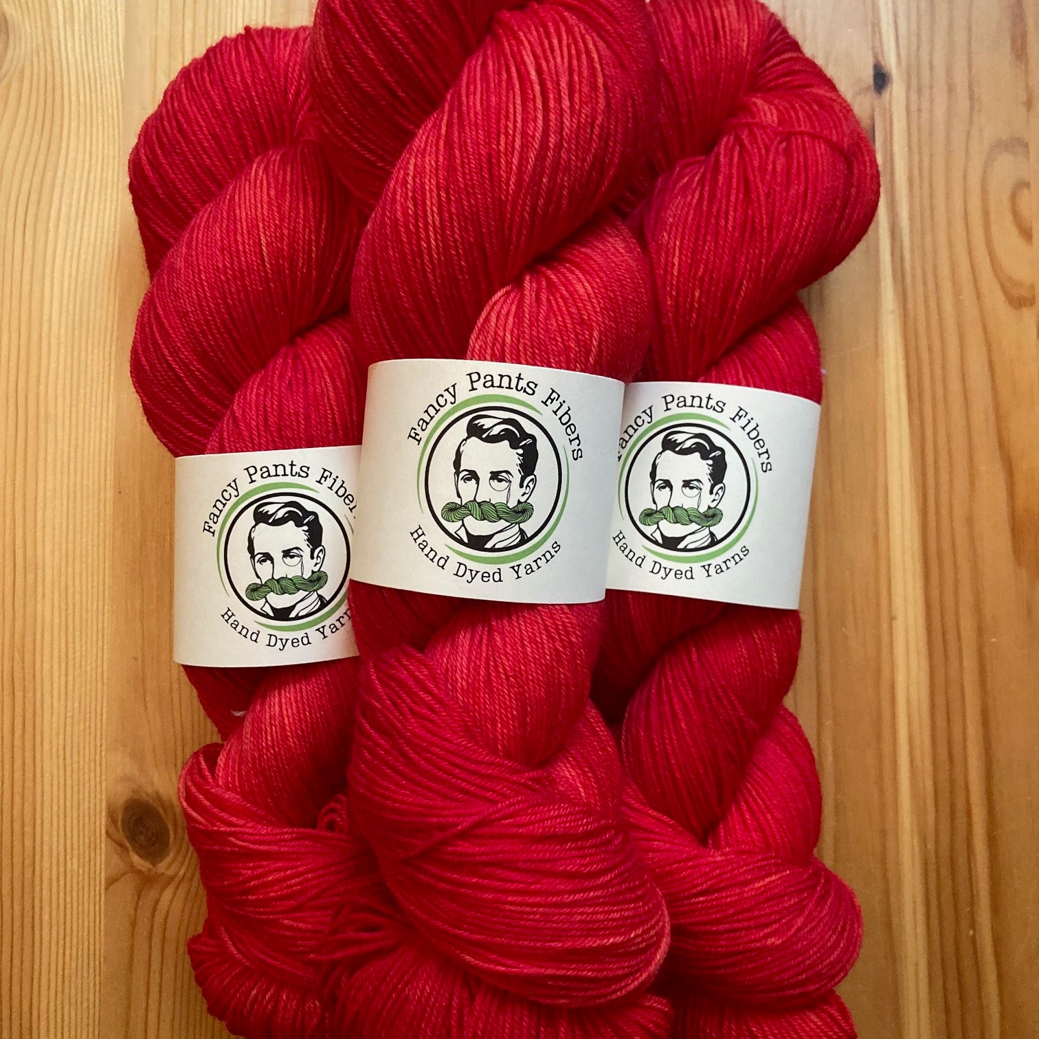 Red yarn skeins with 'Fancy Pants Fibers' labels on a wooden surface